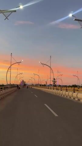 This media file depicts a sunset over a bridge in Meureudu, Indonesia. The sky is painted with vibrant hues of orange, pink, and blue, indicating the time of day is dusk. The bridge itself is a prominent subject, featuring a paved road with white lane markings. Along the sides of the road are decorative lampposts with curved metal supports, illuminating the scene with bright white lights. The bridge is lined with a yellow and white barrier.

Several people are visible on the bridge, some riding motorcycles and others standing along the railing. Their presence suggests the bridge is a place of transit and potentially a scenic spot for onlookers. The surrounding landscape appears relatively flat with some trees and buildings visible in the distance. The overall mood conveyed is peaceful and serene, amplified by the beautiful natural light of the sunset and the calming presence of the people. There is no visible text in the media file.