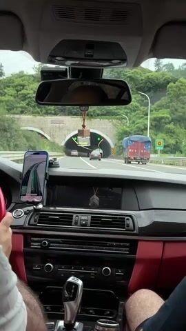 The video shows the interior of a car driving through a tunnel. The perspective is from the passenger seat, looking forward. The dashboard features a GPS navigation system on a smartphone, displaying a route with traffic information. The car's infotainment system is also visible, showing song titles and playback information. The interior of the car has red leather accents.

Outside the car, the scene transitions from a green, tree-lined exterior to the dimly lit interior of a tunnel. The tunnel is illuminated by rows of lights along its ceiling. In the distance, other vehicles can be seen, including cars and a truck. The tunnel appears to be an express or highway tunnel.

There are two visible subjects, likely the driver and passenger, though only their legs and arms are clearly in frame. The overall mood is one of transit and travel. The time of day appears to be daytime, judging by the light outside the tunnel and the green foliage visible. No specific weather conditions are discernible, but the presence of foliage suggests it is not inclement.

Visible text includes "60" on the GPS display, likely indicating a speed limit or current speed, and "740 m" on a sign outside the tunnel, indicating remaining distance. The car's climate control displays "16.0" which could be the temperature setting. The infotainment screen displays song titles, such as "Unlocks and", and artist names like "Ma Jiaming". The location context of Fuyang, China, is provided, which helps to contextualize the setting.