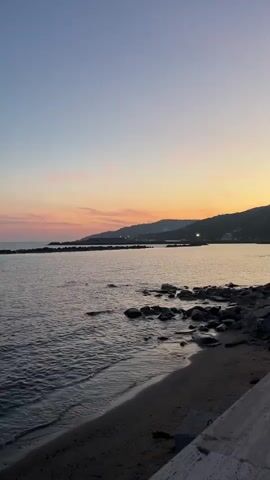 The video depicts a serene coastal scene at dusk. The sky is painted with hues of orange and pink, transitioning to a pale blue as it ascends. A gentle body of water stretches towards the horizon, reflecting the fading light. A small stretch of sandy beach meets the water's edge, interspersed with dark rocks. In the distance, a silhouetted landmass is visible, dotted with faint lights, suggesting a settlement.