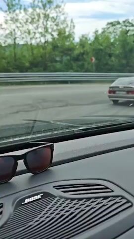 The video shows a car driving on a winding road. The camera is positioned inside another vehicle, showing the dashboard with a pair of sunglasses and a Bose speaker. Through the windshield, we see the road ahead and a car in front of the camera vehicle. The landscape includes lush green hills and trees on either side of the road. The sky is partially cloudy, with patches of blue visible. The car in front of the camera car is silver. The road has white lane markings and red and white signs.