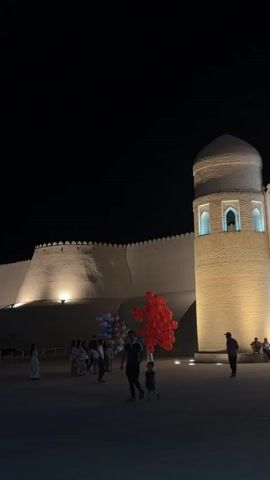 The video shows a nighttime scene with a large, illuminated fortress wall and a tower. A group of people are gathered in front of the fortress, some holding bunches of balloons. One person, presumably a vendor, holds a large cluster of red balloons. The ground is paved, and several figures are visible walking or standing. The overall atmosphere appears festive.