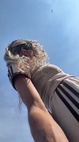 The video begins with a close-up shot of a woman with long, curly blonde hair and sunglasses, looking up at the sky. The camera then pans out to show her on a sandy beach next to a body of water. She is wearing a light-colored t-shirt with brown stripes on the sleeves and collar, and dark pants with stripes down the legs. In the background, there are trees and a few other people walking along the shoreline. The camera continues to pan, showing more of the beach and the water. The woman smiles at the camera throughout the video. The sky is blue with some clouds.