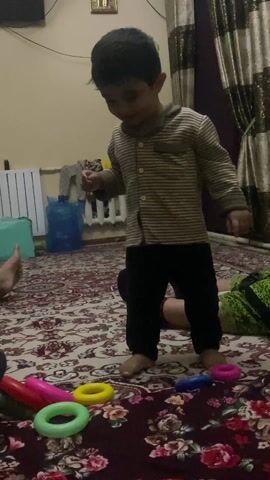 The video shows a young boy playing in a room. The room is carpeted with a colorful floral pattern. There are several people sitting on the carpet, and the boy is standing near them. He is wearing a striped shirt and black pants. There are several colorful toy rings scattered around him. The boy is looking down and appears to be smiling. One of the people sitting on the carpet is pointing at the toys. The background of the room includes a radiator, a curtain, and a television.