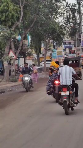 The video shows a rear view of two people riding on a motorcycle on a paved road in what appears to be a town or city. The rider, wearing a checkered shirt, is operating the motorcycle. The passenger behind them is wearing a traditional Indian outfit, a sari, and a yellow headscarf. The motorcycle's brake light is illuminated. In the background, there are buildings lining the street with various signs and advertisements, as well as other motorcycles and vehicles parked along the side. Trees with green foliage are visible. The road surface is dusty and appears to be in moderate condition. The scene suggests a typical street view in an urban environment in India.