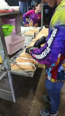 A person in a multicolored long-sleeved shirt and jeans is making hotdogs at an outdoor stand. They are wearing black gloves and placing hotdogs into buns that already have mustard on them. There is a tub of hotdogs to their left and a green strainer behind them. Another person in a pink and purple shirt is visible in the background. The ground appears to be concrete. Loud music is playing in the background.