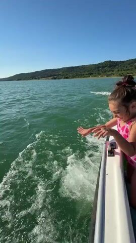A young girl in a pink swimsuit with white hearts is on a boat. She is smiling and looking towards the camera. The boat is moving through green water, creating a wake. The background shows a body of water and a tree-covered hillside under a clear blue sky.
