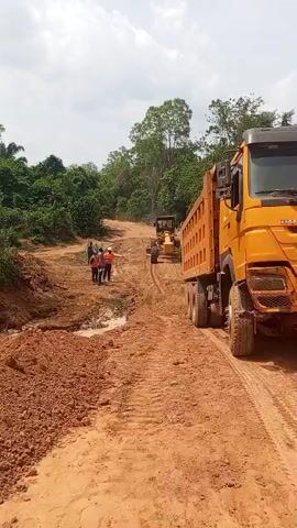 This video depicts an active construction site in Aloko, Nigeria, under a cloudy sky during the daytime. The scene is dominated by reddish-brown earth, characteristic of the region, with dense green foliage bordering the area.

Several men are present on the unpaved dirt road, some wearing safety vests and hard hats, suggesting they are construction workers or supervisors. They are gathered near a muddy puddle and appear to be in a discussion, with one man pointing towards the ground, possibly giving instructions or inspecting the work.

Heavy machinery is also visible: a large yellow motor grader is positioned further down the road, and a yellow dump truck is parked or moving slowly on the right side of the frame. Piles of excavated or moved soil are present on the left. The overall activity suggests road construction or land development is underway. No discernible text is visible in the video.