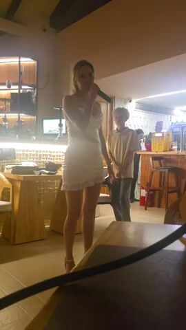 A woman in a white dress stands with a microphone in her hand, singing. She is in a dimly lit room that appears to be a bar or restaurant. In the background, a person stands behind a counter with bottles of drinks on shelves. A fire extinguisher is visible on the wall.