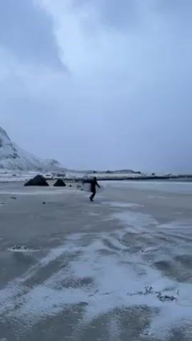 A person in a wetsuit carries a surfboard across a snow-dusted beach towards the water. The sky is overcast and a grey color. The ocean is choppy with small waves. Some other surfers are already in the water. Snow dusts the sand. In the distance, a small hill covered with snow is visible.