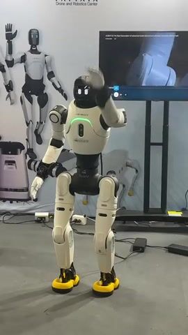 The video shows a humanoid robot with a white and black body and yellow feet. The robot is standing on a gray floor in front of a wall with a banner that says "PATTAYA Drone and Robotics Center". There are other robots visible in the background. The robot in the foreground raises its right arm and waves. It then lowers its arm and bends its knees. The robot's head has two glowing eyes.