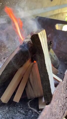 The video shows a close-up view of a small campfire being built inside a metal fire pit. Several pieces of wood are arranged in a teepee formation, with smaller pieces at the base and larger pieces stacked on top. Flames are visible, licking up from the center of the woodpile, and smoke billows around the flames. The fire pit appears to be made of rusted metal and is sitting on a bed of ashes. The background is blurred but suggests an outdoor setting with greenery. The camera slowly zooms out to reveal more of the fire pit and its surroundings.