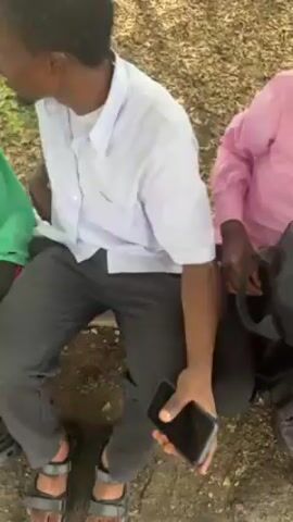 The video shows several young people in casual clothing sitting together outside on a stone or concrete structure, probably a curb or low wall. One is holding a smartphone. Another has a twig or stick in his hand, which he hands to the person next to him, who then begins to play with it, laughing. The environment includes trees and grass, and other people can be seen in the background.