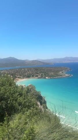 The video shows a panoramic view of a tropical beach from a high vantage point. The water is a clear turquoise, with small waves breaking on the sandy shore. Several people are visible in the water and on the beach. In the distance, there are hills covered in green vegetation and the blue expanse of the ocean stretches to the horizon. The sky is clear and blue. There are boats in the water. The video pans slowly across the scene, offering different perspectives of the beach and the surrounding landscape.