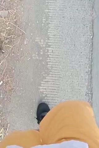 A first-person perspective video shows a person walking on a paved surface. The person is wearing black shoes and orange shorts. To the left of the path, there is dry vegetation and scattered debris. The paved surface has a textured pattern, possibly from the way it was laid. The video captures the rhythmic motion of the person's feet as they walk forward.