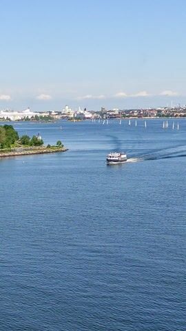 This is a high-angle, sideways video clip of a boat traveling on a large body of water, with a cityscape visible on the left side of the frame. The water is a deep blue and appears calm, with a visible wake trailing behind the boat. The boat itself is white with dark trim and appears to be a passenger vessel. The cityscape consists of numerous buildings of varying heights and architectural styles, some appearing to be industrial or commercial, and others resembling residential or office buildings. There are also trees and greenery along the shoreline, suggesting a park or natural area adjacent to the urban environment. The sky is a clear, light blue, with a few scattered white clouds, indicating fair weather. The overall impression is that of a sunny day, likely in the late morning or early afternoon, given the brightness of the light. The location is identified as Helsinki, Finland, which aligns with the visual cues of a coastal city with a significant harbor or waterfront area. No people are clearly discernible on the boat or in the cityscape, and no text is visible within the frame.