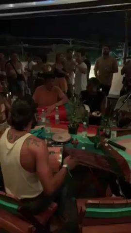 A video shows a group of people gathered around a table at night, enjoying a musical performance. Several individuals are playing instruments, including percussion and a small guitar-like instrument, while others clap and sing along. The musicians are seated at a table covered with a colorful patterned cloth. Beer bottles, water bottles, and small plants are arranged on the table. A microphone stands in the center, indicating that the performance is amplified. The crowd consists of men and women of varying ages, most of whom appear to be standing or dancing. The setting is an outdoor venue, possibly a patio or deck, with dim lighting.