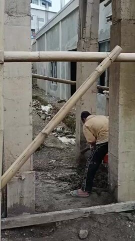 A man is using a hoe to dig in a muddy area between concrete walls. The walls appear to be part of a building under construction, with bamboo support structures visible. The man is wearing a tan shirt, dark pants with a light stripe down the side, and slides. In the background, there's another building and some rubble.