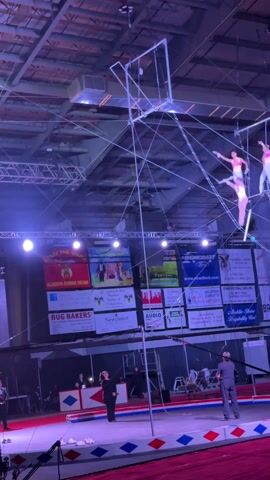 The video shows two trapeze artists performing in a circus tent. One artist in a pink and white outfit hangs from a trapeze while the other, in a white and pink outfit, hangs above them. The artist on the lower trapeze releases their grip and swings forward, making a flip in the air before being caught by the other artist. In the background, a large screen displays the circus' logo, and several people stand watching below. The lighting is bright, with colorful spotlights illuminating the performance area.