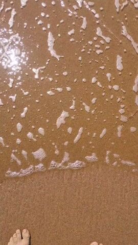 The video shows waves washing onto a sandy beach. The sand appears to be a light brown color, and the water creates white foam as it moves across the sand. There are some reflections on the water, suggesting sunlight. The waves are not large, and the overall scene is calm and peaceful. The camera is positioned close to the ground, offering a close-up view of the interaction between the waves and the sand.