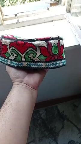 A hand holds up a traditional embroidered cap, displaying its colorful floral pattern. The cap is predominantly white with red and green floral motifs and a blue border. The background shows a sunlit window with greenery visible outside and a dark, patterned surface below.