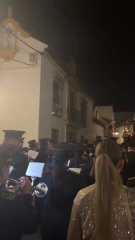 At night, a group of musicians dressed in black uniforms with caps plays instruments, likely brass, in a narrow street lined with white buildings. They are playing from sheet music placed on stands in front of them. A woman with a blonde ponytail and wearing a shimmering gold top stands in front of the group. Further down the street, a religious figure or scene atop a platform adorned with lights is visible. The scene appears to be a religious procession or parade. The sky is dark.