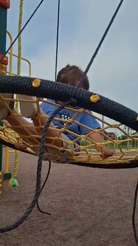 A young girl with brown hair in a ponytail, wearing a blue t-shirt and white socks, sits on a circular net swing on a playground. The swing is suspended by four black ropes. The girl is holding onto the net with her hands and her legs are bent at the knees. She appears to be looking down at the net. The ground beneath the swing is covered with small, reddish-brown gravel. In the background, there are some yellow and green playground structures, a fence, some buildings, and a cloudy sky. The video is shot from a low angle, looking up at the girl on the swing.