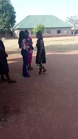 Four women stand in a group in front of a building with a green roof. One woman is wearing a black dress, one is wearing a pink dress, one is wearing a patterned shirt and blue pants, and the fourth is wearing a black dress with white polka dots. The ground is dirt. A large tree is in the background. The women appear to be talking.