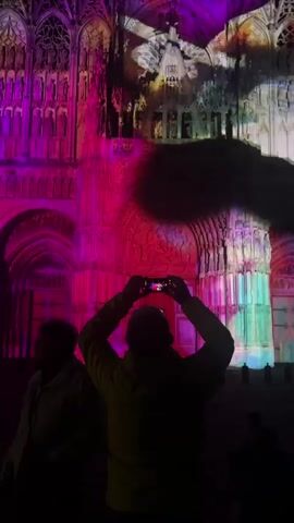 Two people stand in the foreground watching a light show projected onto a cathedral. The person in the right foreground is silhouetted and holding a smartphone above their head, recording the event. The cathedral's facade is illuminated with various colors and moving patterns, including a dark, swirling shape that appears to mimic a dragon or a creature. The light show transitions from purple and pink hues to a display of red, white, and blue, resembling the French flag. The person in the left foreground is also silhouetted and appears to be observing the light show.