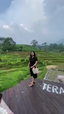A woman wearing a black dress and sandals stands on a wooden platform overlooking a tiered rice field. She is holding a small white purse and looking out at the scenery. To her left, a child sits on a cushioned bench. Behind the woman, there is a hammock-style seating area. The environment is lush and green, with trees dotting the landscape under a partly cloudy sky.