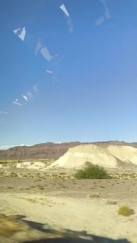 The video shows a view from a vehicle moving through a desert landscape. The sky is clear and blue. In the middle ground, there are rolling hills and a large mound of pale earth. In the background, there are mountains with reddish-brown rock formations. The foreground shows a dusty road or path, with some dry vegetation. The overall impression is one of arid, open country.