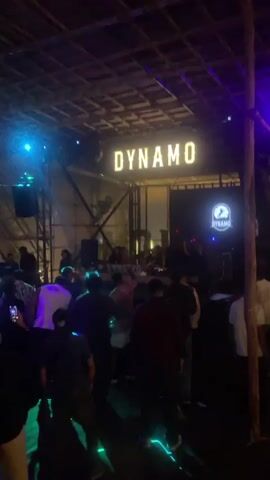 A crowd of people is gathered inside a dimly lit venue. The ceiling appears to be made of wooden beams and a woven material. A large illuminated sign spelling "DYNAMO" hangs from the ceiling. Below it, another sign with a circular logo that also says "DYNAMO" is visible. Blue and purple lights are casting a glow on the scene, and some areas are illuminated by spotlights creating beams of light. People are standing and some appear to be dancing. The overall atmosphere suggests a nightlife or entertainment venue.