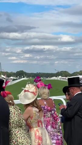 The video shows a horse race from the perspective of spectators standing behind a white railing. The spectators are dressed in formal attire, including top hats and elaborate hats. The race track is a lush green field, and there are trees and buildings in the distance. The sky is blue with scattered clouds. The video is shot from a low angle, looking up at the spectators and down at the race track. The video is shaky, suggesting it was filmed handheld. The sound of the crowd and the horses can be heard faintly in the background.