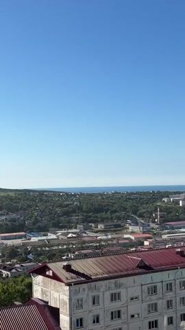 A panoramic view of a town is shown from an elevated position. The sky is clear and blue. In the foreground, the rooftops of several buildings are visible, one with a red, corrugated metal roof and another with a lighter-colored facade. Beyond the buildings, the town spreads out with a mix of residential houses, industrial buildings, and smaller structures, all nestled amongst trees. In the distance, the horizon meets the sea under a hazy sky. The camera pans slowly across the landscape.