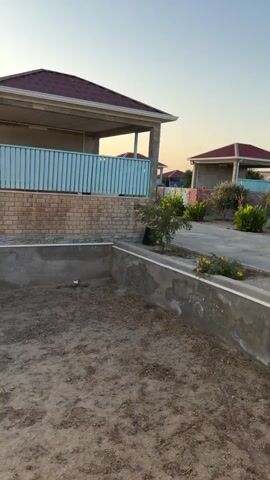 The video shows a view of a beach resort area. The first shot shows a newly constructed building with a red roof and brick walls, and a light blue fence. The area in front of the building is dirt and appears to be under construction or preparation for a pool. The second and third shots show a sunset over the ocean. Two young people, appearing to be children or teenagers, are walking on a path in the distance. The fourth and fifth shots show a view of the beach with several small structures, possibly changing rooms or cabanas, and young olive trees planted in rows. The sky is clear and the sun is low on the horizon, indicating it is either sunrise or sunset. The overall impression is of a developing resort area with a scenic coastal location.