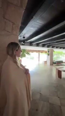 A woman in a tan shawl is standing inside a building with stone walls and looking out at a tropical rainstorm. Another person in a white shirt and blue cap walks behind her. The room she is standing in has a dark wood beam ceiling and a tile floor.  The rain is falling heavily on a tiled outdoor area with palm trees and tropical plants.