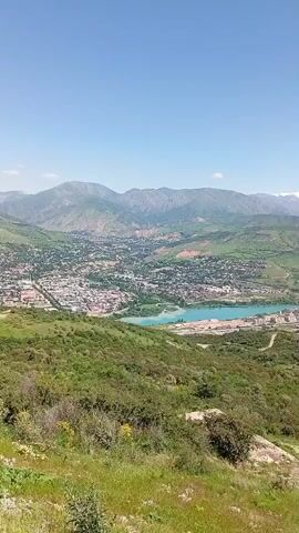 The video shows a panoramic view of a landscape with a town, a lake, and mountains in the background. The town is nestled in a valley, surrounded by green hills and trees. A turquoise lake is adjacent to the town, reflecting the clear blue sky. In the distance, mountains can be seen, some with snow-capped peaks. The foreground is filled with green vegetation, including grass, bushes, and small trees. The overall impression is one of natural beauty and tranquility.