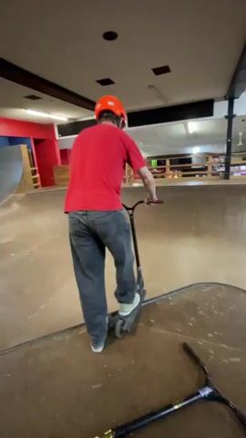 The video shows a skatepark with two individuals on scooters. One person, wearing a red shirt and orange helmet, is seen riding a scooter inside a bowl-shaped area. The other person, wearing a red shirt and orange helmet, rides a scooter in the bowl. The person in the red shirt and orange helmet performs a trick, flipping in the air. The video ends with a close-up of the scooter handlebars.