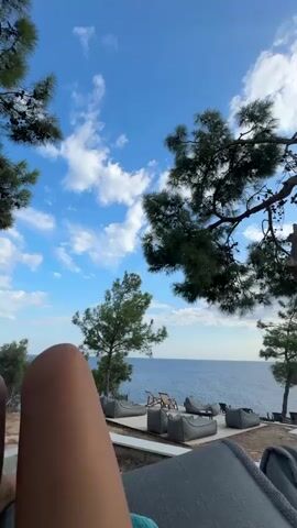 This video shows an outdoor seating area overlooking a calm blue sea. There are several large, grey beanbag chairs and some lounge chairs arranged on a wooden deck. Pine trees are scattered around the area, with one large tree in the foreground partially obscuring the view. The sky is a bright blue with scattered white clouds. In the background, a person is walking towards an outdoor grill area with a white tiled roof. The overall scene suggests a relaxed, coastal atmosphere, likely a resort or a cafe by the sea.