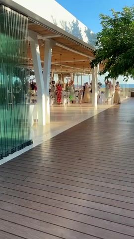 A group of people are gathered at an outdoor event, possibly a wedding reception or a celebration, in Peyia, Cyprus. The scene is set on a wooden deck overlooking a building with a white roof and white pillars, featuring large glass doors that offer a glimpse into the interior. Inside, tables are set up, suggesting a dining or reception area. Several individuals are visible inside, some appearing to be dancing or raising their arms in celebration. The atmosphere seems festive and joyful.

The time of day appears to be late afternoon or early evening, as indicated by the warm sunlight casting long shadows and illuminating the scene with a golden hue. The sky is clear and blue, suggesting good weather. A lush green tree is prominent on the right side of the frame, providing shade and a natural element to the setting. In the background, a hint of the sea or a large body of water can be seen, along with a hazy horizon, typical of a coastal location.

The people are dressed in celebratory attire. One woman in the foreground on the right is wearing a light-colored, flowing dress and appears to be dancing or walking with a graceful stride. Inside, there are people in colorful dresses and suits, further emphasizing the festive nature of the gathering. The overall impression is one of a happy occasion taking place in a beautiful, open-air venue with a view. There is no visible text in the media file.