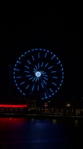 This video shows a Ferris wheel at night. The Ferris wheel is lit with blue and purple lights that change color. The Ferris wheel is in front of a dark sky, with some buildings and lights visible below. The water in front of the buildings reflects the lights.