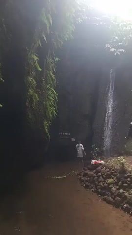 The video shows a narrow canyon with lush greenery on the left wall and a waterfall cascading down the right wall. The bottom of the canyon is filled with a shallow, muddy river. Two people are visible in the scene. One person, wearing a white shirt and shorts, is standing in the river facing away from the camera, with their back to the waterfall. Another person, wearing a dark shirt and shorts, is standing on the right bank of the river, also facing away from the camera, near the base of the waterfall. The environment appears to be natural and possibly remote. The lighting suggests it might be overcast or the sun is positioned such that it creates a bright, hazy effect at the top of the canyon. The overall impression is of a serene and secluded natural spot.