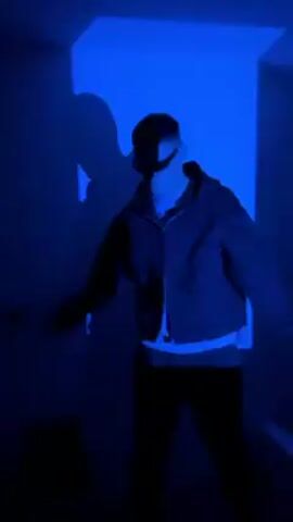 A person wearing a black jacket, white shirt, and sunglasses stands in a dark room illuminated by blue and purple lighting. The person is slightly out of focus and their shadow is cast against a white wall. The person is moving their arms. The background is dark and the walls are plain. Music is playing in the background.