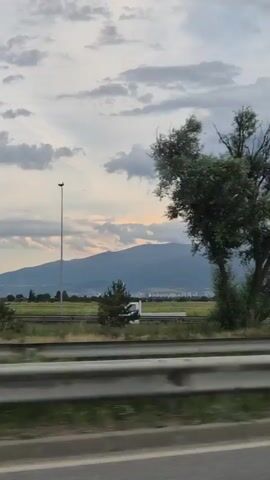 A side view of a highway with traffic in motion. In the foreground, a metallic guardrail runs along the edge of the road, bordered by green grass. Beyond the guardrail, a flat, grassy field stretches towards a distant mountain range. A few trees are visible on the left side of the frame, with one notably bare tree. The sky is overcast with dramatic, dark clouds, but a hint of sunset light can be seen through them. Several vehicles are captured in motion on the highway: a white station wagon passes by, followed by a yellow van and a dark-colored car. In the distance, the mountains are shrouded in a hazy atmosphere, with a few buildings visible at their base. The overall impression is of a journey on a cloudy day with a scenic, expansive landscape.