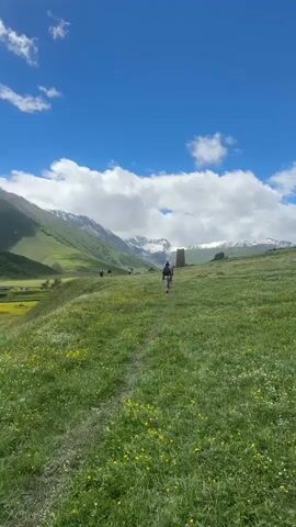The video shows a group of people walking on a grassy hill under a bright blue sky with scattered white clouds. In the background, there are majestic mountains, some of which are snow-capped. A stone tower stands on the hill in the distance. The grass is green and dotted with small yellow and white flowers. The people are walking away from the camera, following a path through the grass. One person in the foreground is wearing a backpack. The overall atmosphere is serene and picturesque, suggesting a peaceful outdoor excursion.