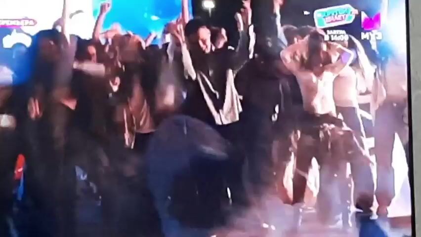 The video shows a crowd of people dancing at what appears to be an outdoor event or concert. The lighting is dim with some bright spotlights creating a festive atmosphere. In the foreground, a man in a striped shirt is dancing, while to his right, a woman in a white top is also enthusiastically dancing. Other people are visible in the background, many with their hands in the air. The screen also displays graphics, including a logo that says "МУЗ" and "BETTER. SMART." The overall impression is one of a lively party or performance.