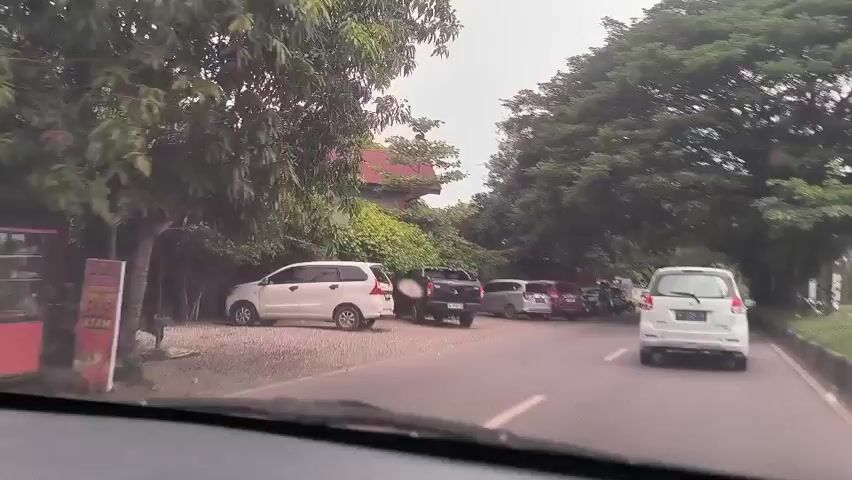 The video shows a view from inside a car driving down a road with trees on either side. Several parked cars, including a white van, a black pickup truck, and a maroon car, line the side of the road. A few people are standing near the parked cars, including one wearing a headscarf. A covered area with tables and chairs is visible behind the cars, suggesting a potential gathering place. The sky appears overcast.