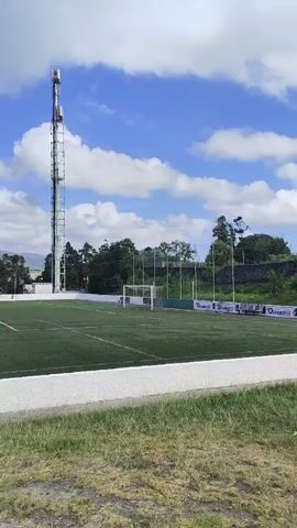 The video shows an outdoor view of a soccer field with artificial turf. A tall telecommunications tower stands on the left side of the frame. There are soccer goals on the field. In the background, there are trees, buildings, and hills under a blue sky with white clouds. The video pans slightly to the right.