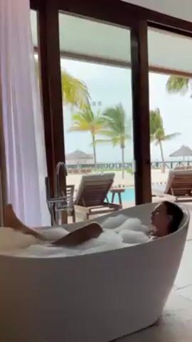 The scene is a woman enjoying a bubble bath in a freestanding white bathtub, situated inside a room with a view of a tropical beach. Palm trees are visible through the glass doors, along with beach chairs, a pool, and a pier. The woman is lying back in the tub, her legs slightly raised, appearing relaxed. The bathroom has white curtains and wooden door frames. The overall ambiance is serene and luxurious.