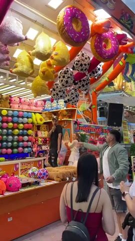 The video shows a fair booth with a variety of plush prizes hanging from the ceiling, including donuts, chickens, pigs, and dalmation-print balls. In front of the prizes is a wall of colorful balloons. A man in a black shirt and ripped jeans stands behind the booth, while other people gather around the booth's counter. A sign on the booth says “Rompiendo.”