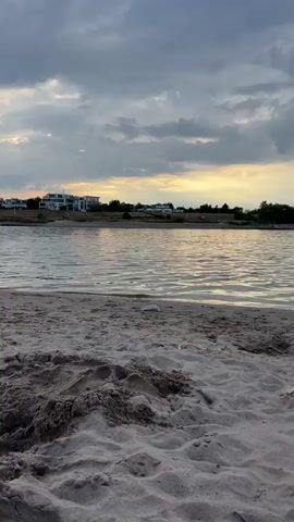 The video shows a close-up view of sandy beach in the foreground. In the background, there is a body of water, possibly a lake or river. Beyond the water, there is a shoreline with trees and buildings. The sky is cloudy with some patches of sunlight breaking through. The camera perspective is low to the ground, suggesting it is positioned on the sand. At 00:03, a person's bare foot emerges from the sand, indicating they are buried or resting in the sand. The overall scene depicts a peaceful, natural setting at dusk or dawn.